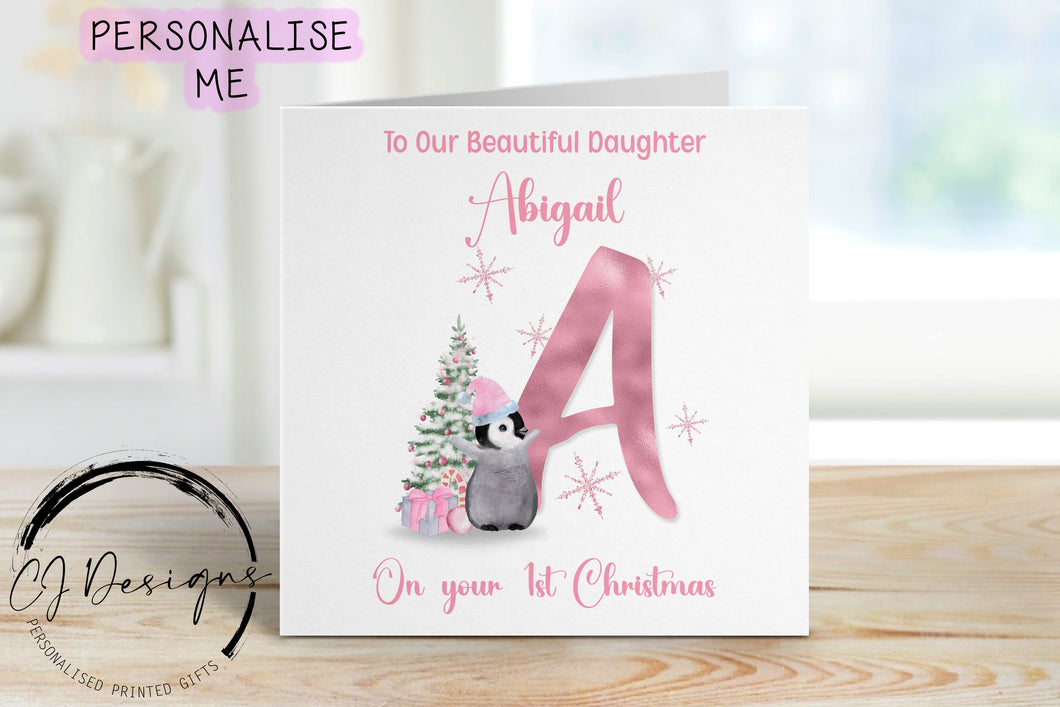 Daughter First Christmas christmas card in a pink theme with picture of baby penguin wearing a pink hat in front of a christmas tree and next to a large letter which depicts the first letter of the childs name.