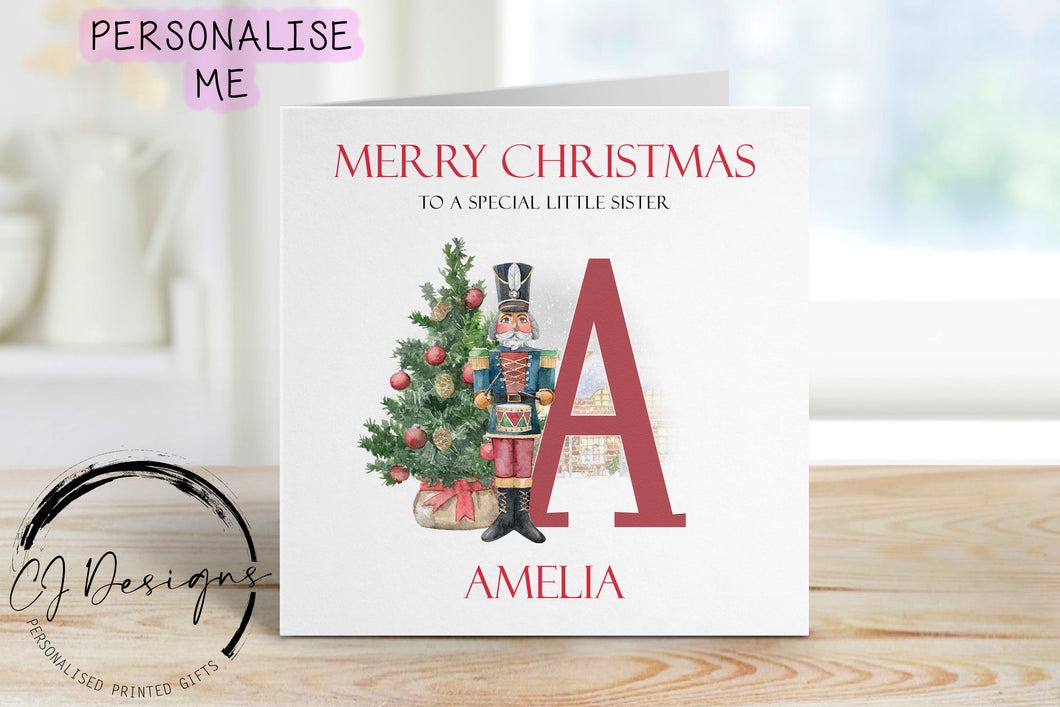 Little Sister personalised Christmas card with a nutcracker with Christmas Tree sstood next to a large red letter which depicts the first letter of your little